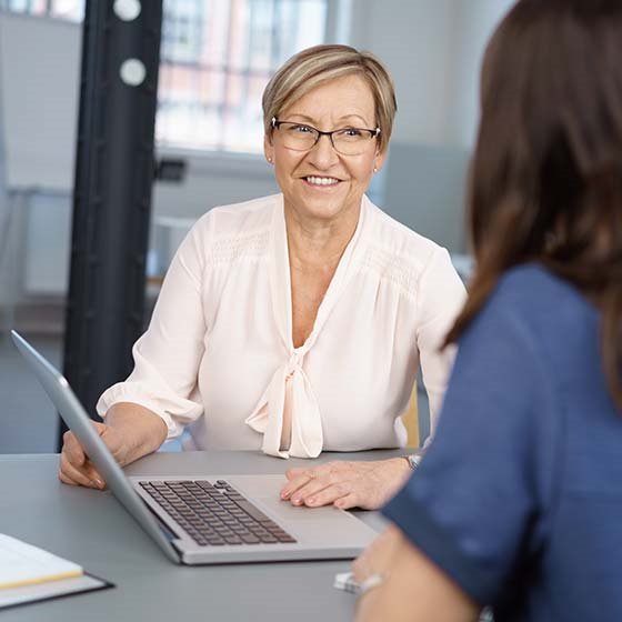 Two businesswoman discussing a work project seated together at a table in front of a laptop computer with focus to a middle-aged lady