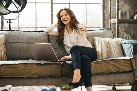 Photo of happy woman in elegant clothing sitting on couch in front open laptop computer in loft living room