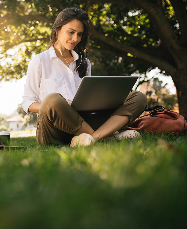 Eine junge Frau sitzt in der Wiese und schaut auf ihren Laptop