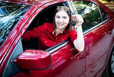 MÃ¤dchen sitzt hinter dem Steuer eines roten Autos und hÃ¤lt stolz den AutoschlÃ¼ssel in der Hand