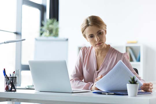 Eine Frau sitzt vor dem Laptop und schaut in Unterlagen